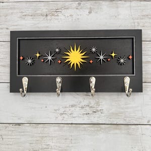 May include: Black wooden key holder with a celestial design. The design features a yellow sun and various star shapes in white, orange, and gray. Four silver hooks are attached below the design for hanging keys or other small items.