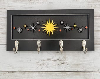 May include: Black wooden key holder with a celestial design. The design features a yellow sun and various star shapes in white, orange, and gray. Four silver hooks are attached below the design for hanging keys or other small items.