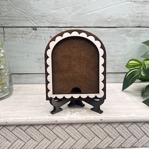 May include: A small, dark brown wooden picture frame with a white scalloped border. The frame has a built-in stand and a slot at the bottom. The frame is empty and ready to display a photo or artwork.