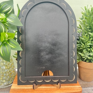 May include: A black wooden arched chalkboard with a scalloped edge, displayed on a wooden stand. The chalkboard is set on a rectangular wooden base. Decorative plants are in the background.