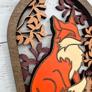 May include: A layered wooden art piece featuring a fox in shades of orange and cream, set against a backdrop of autumn leaves. The fox is the focal point, framed by a dark brown arch and intricate leaf cutouts.