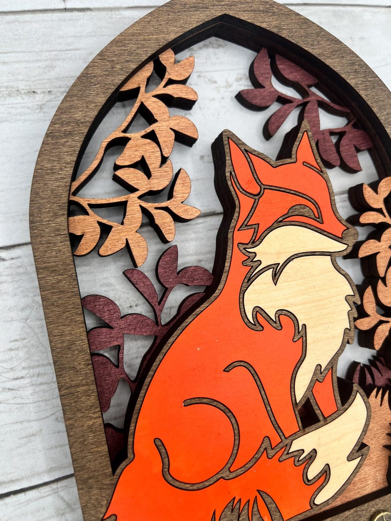 May include: A layered wooden art piece featuring a fox in shades of orange and cream, set against a backdrop of autumn leaves. The fox is the focal point, framed by a dark brown arch and intricate leaf cutouts.
