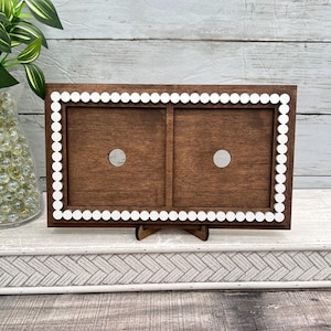 May include: A wooden double photo frame with a dark brown finish. The frame has a white scalloped border and two circular openings. It is displayed on a small wooden stand.