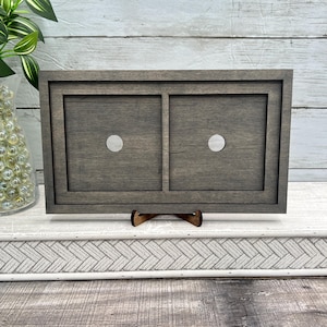 May include: A grey wooden frame with two recessed squares, each with a silver dot. The frame is supported by a small wooden stand. The background features a white wooden surface and a green plant.