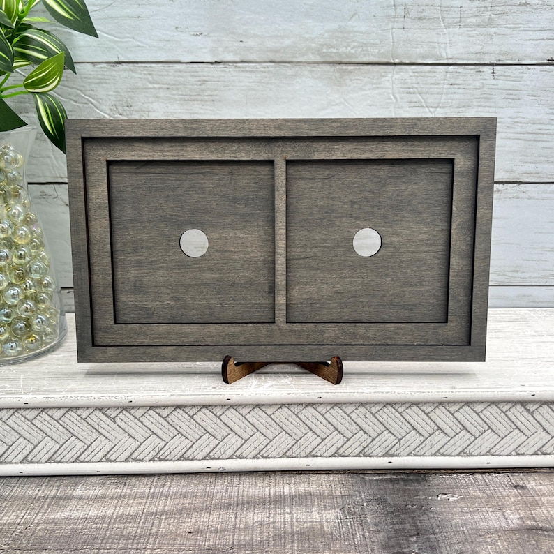 May include: A grey wooden frame with two recessed squares, each with a silver dot. The frame is supported by a small wooden stand. The background features a white wooden surface and a green plant.