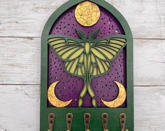 May include: A decorative wooden key holder featuring a moth design. The key holder has a green frame with a purple background and a yellow moth. The design includes a moon and crescent moons. It has five bronze hooks.