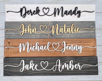 May include: Four wooden signs with names connected by a heart symbol. The top sign has "Derek & Mandy" in black script on a white background. The second sign has "John & Natalie" in gold script on a gray background. The third sign has "Michael & Jenny" in white script on a brown background. The bottom sign has "Jake & Amber" in white script on a dark gray background.