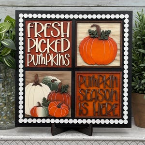 May include: A square wooden sign with a black frame and white bead border, divided into four quadrants. The top left reads "FRESH PICKED PUMPKINS" in white letters. The top right shows an orange pumpkin. The bottom left features four pumpkins, and the bottom right reads "PUMPKIN SEASON IS HERE" in dark brown letters.