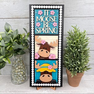 May include: A wooden sign with a black frame features the phrase "IT MOOSE BE SPRING" above two moose illustrations. The moose are decorated with pink flowers and a yellow hat. The sign is propped up on a wooden stand.