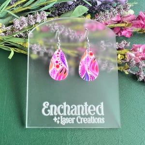 May include: A pair of teardrop-shaped earrings with a vibrant pink, orange, and purple pattern. The earrings are displayed on a clear acrylic stand with the text "Enchanted Laser Creations". The earrings have silver-colored hooks.