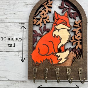 May include: Wooden key holder featuring a fox design. The fox is orange and white, set against a backdrop of autumn leaves. The key holder is 10 inches tall and 6.5 inches wide, with five bronze-colored hooks.