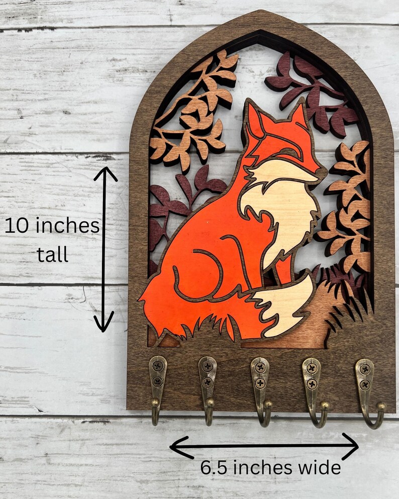 May include: Wooden key holder featuring a fox design. The fox is orange and white, set against a backdrop of autumn leaves. The key holder is 10 inches tall and 6.5 inches wide, with five bronze-colored hooks.