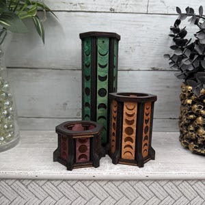 May include: Three hexagonal wooden containers of varying heights featuring laser-cut moon phase designs. The tallest container is green, the medium is orange, and the shortest is purple. Each piece has dark brown structural frames and open tops.