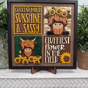 May include: A framed wooden art piece with four square panels. Each panel features a Highland cow design with sunflowers and text that reads "Current Mood Sunshine & Sassy" and "Fluffiest Flower in the Field".