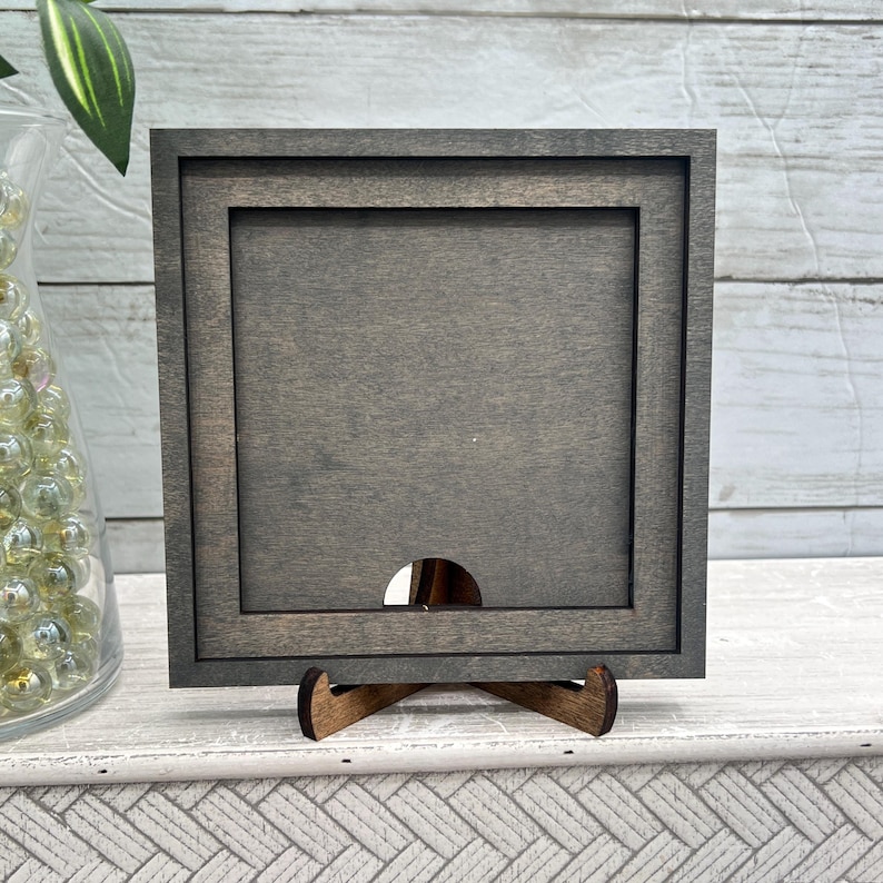 May include: A square, grey wooden picture frame with a dark stain, supported by a small wooden easel. The frame features a recessed inner border and a circular cutout at the base. The background is a white wooden surface.