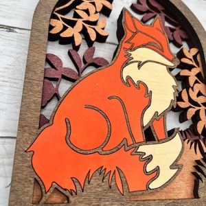 May include: Wooden key holder featuring a layered design of a fox in shades of orange, cream, and brown. The fox is set against a backdrop of stylized leaves in a dark brown frame. Includes four brass key hooks.