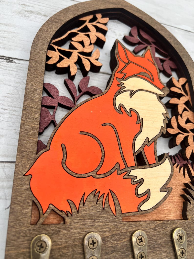 May include: Wooden key holder featuring a layered design of a fox in shades of orange, cream, and brown. The fox is set against a backdrop of stylized leaves in a dark brown frame. Includes four brass key hooks.