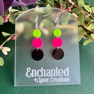 May include: A pair of dangle earrings with a neon green, hot pink, and black circular design. The earrings are suspended from silver-colored hooks. The earrings are displayed on a clear, frosted acrylic stand with the text "Enchanted Laser Creations".