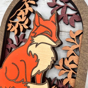 May include: A layered wooden fox art piece. The fox is orange and cream-colored, set against a backdrop of fall foliage in shades of brown and burgundy. The artwork is framed within a dark brown arch.