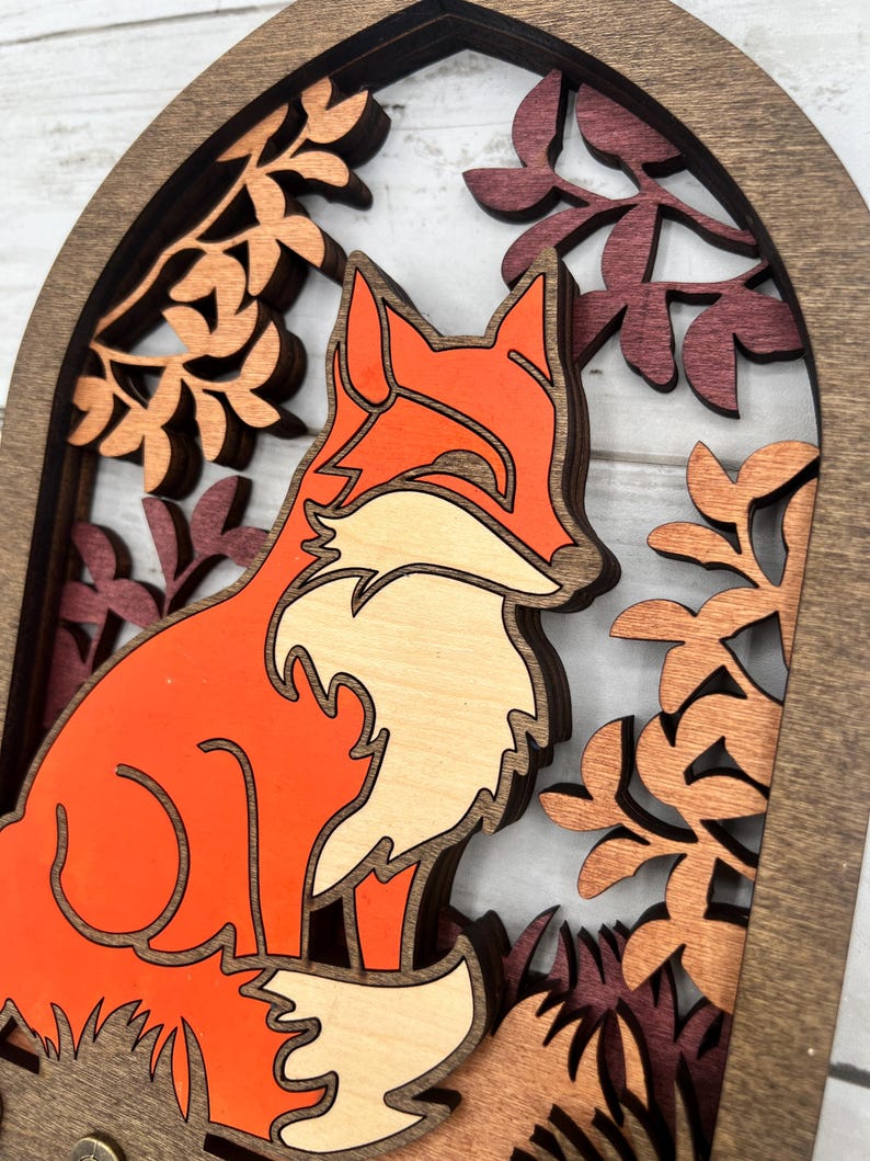 May include: A layered wooden fox art piece. The fox is orange and cream-colored, set against a backdrop of fall foliage in shades of brown and burgundy. The artwork is framed within a dark brown arch.