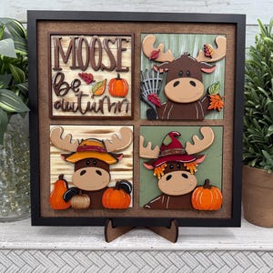 May include: A framed wooden art piece featuring four panels with a fall theme. Each panel showcases a cartoon moose with autumn accessories like hats, pumpkins, and leaves. The top left panel has the text "MOOSE be autumn".