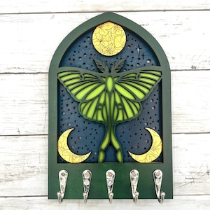 May include: A green and blue wooden key holder featuring a layered design of a moth, moon, and stars. The moth is lime green, and the moon is yellow. The key holder has five silver hooks.