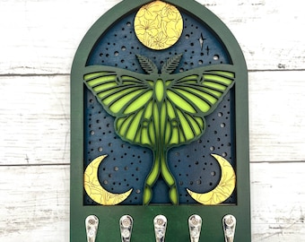 May include: A green and blue wooden key holder featuring a layered design of a moth, moon, and stars. The moth is lime green, and the moon is yellow. The key holder has five silver hooks.