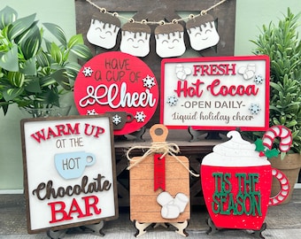 May include: A collection of festive wooden signs and decorations with a winter holiday theme. The signs feature phrases like "Fresh Hot Cocoa" and "Warm Up at the Chocolate Bar" in red and white. There is also a wooden cutting board with marshmallows.