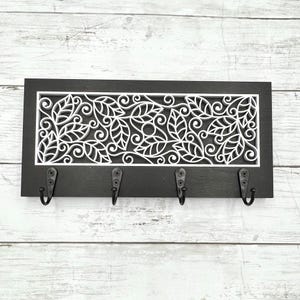 May include: A black wooden wall-mounted key holder with four metal hooks. The key holder features a white, laser-cut leaf and vine design. The key holder is rectangular and is mounted on a white wooden wall.