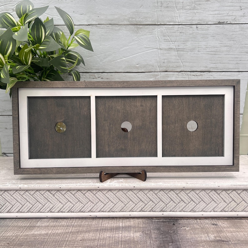 May include: A wooden display frame with three square openings, each with a circular hole. The frame is dark grey with a white border. One opening has a gold-coloured accent. The frame is supported by a small wooden stand.