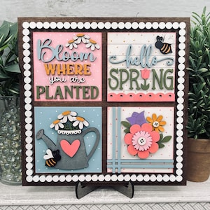 May include: A wooden spring-themed sign with four square panels. Each panel features floral designs, bees, and phrases like "Bloom Where You Are Planted" and "hello SPRING". The sign is framed with a white dotted border and a brown wooden frame.