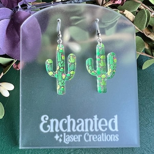 May include: A pair of cactus-shaped earrings with a green and yellow pattern, hanging from silver hooks. The earrings are displayed on a clear acrylic stand with the text "Enchanted Laser Creations".