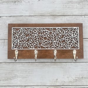 May include: A wooden key holder with a dark brown frame and a white, laser-cut leaf design. Four silver hooks are attached below the decorative panel. The key holder is mounted on a light-colored, wooden plank background.
