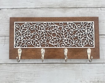 May include: A wooden key holder with a dark brown frame and a white, laser-cut leaf design. Four silver hooks are attached below the decorative panel. The key holder is mounted on a light-colored, wooden plank background.