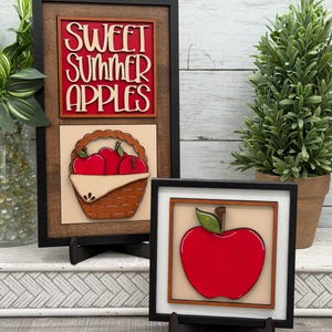May include: Two framed wooden signs with black borders. The taller sign features the text "SWEET SUMMER APPLES" in cream lettering on a red background above a basket of red apples. The smaller square sign displays a single large red apple.