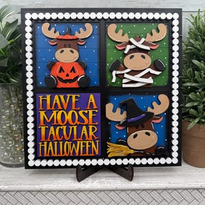 May include: A square Halloween-themed wooden sign with four panels. Each panel features a cartoon moose in a costume. The sign has a black frame with a white dotted border. The text "HAVE A MOOSE TACULAR HALLOWEEN" is in the center.