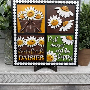 May include: A decorative wooden sign featuring a black frame with white polka dots. The sign is divided into four squares, each with a daisy design. The bottom left square reads "Farm Fresh Daisies". The bottom right square reads "Pick daisies and be happy".