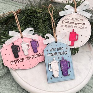 May include: Wooden cup-themed ornaments in pink, blue, and white, each with a different phrase. The pink ornament says "Obsessive Cup Disorder," the blue one says "Work Hard, Hydrate Harder," and the white one says "Just Me and My Emotional Support Cup Against the World."