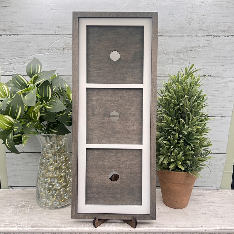 May include: A grey wooden frame with three sections, each with a circular opening. The frame has white borders and stands on a small wooden stand. Decorative plants in vases and pots are on either side of the frame.