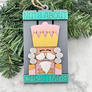 May include: A wooden Christmas ornament featuring a nutcracker design. The ornament has a gray background with a pink nutcracker wearing a gold crown. The text "NUTS ABOUT" and "CHRISTMAS" are in teal.