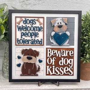 May include: A framed wooden sign with a black border, featuring four squares with dog-themed designs. The top left square reads "dogs welcome people tolerated." Other squares show cartoon dogs and the phrase "Beware of dog kisses."