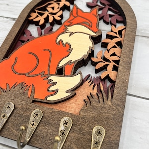 May include: Wooden key holder featuring a layered design of a fox in shades of orange, cream, and brown, set against a backdrop of autumn leaves. Includes four bronze-colored hooks for hanging keys or small items.