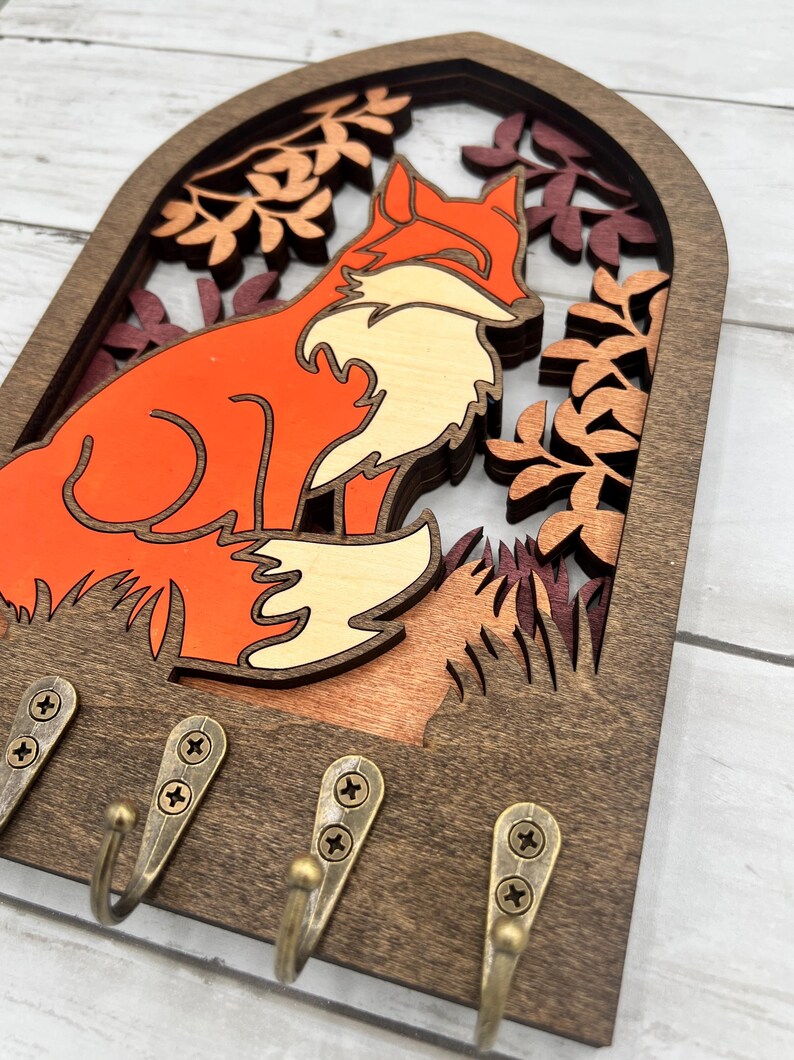 May include: Wooden key holder featuring a layered design of a fox in shades of orange, cream, and brown, set against a backdrop of autumn leaves. Includes four bronze-colored hooks for hanging keys or small items.