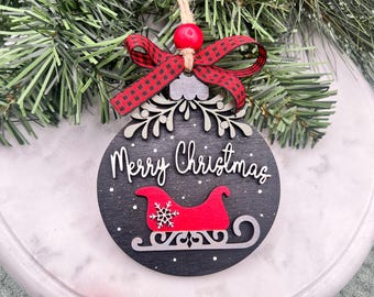 May include: A round, black Christmas ornament with the words "Merry Christmas" in white script. A red sleigh with a snowflake detail is featured below the text. The ornament is decorated with a red and black plaid bow and a red bead.