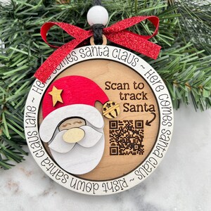 May include: A wooden Christmas ornament featuring a Santa Claus design with a red hat and white beard. The ornament has the text "Here Comes Santa Claus" and "Scan to track Santa". A red glitter ribbon is attached for hanging.