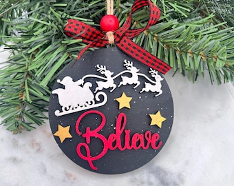 May include: A black Christmas ornament with a Santa and reindeer design, the word "Believe" in red script, and gold stars. It has a red and black plaid ribbon and a jute string for hanging.