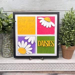 May include: A framed artwork with four colorful squares. Each square features a daisy design and a phrase: "stop and smell the daisies," "bloom," "pick daisies and be happy." The frame is black, and the artwork is displayed on a white surface.