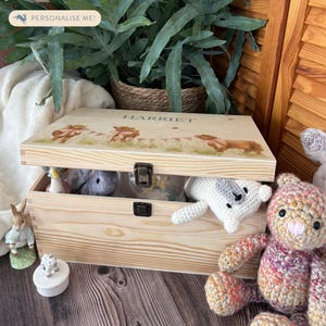 May include: A wooden keepsake box with a personalised name, "HARRIET", on the lid. The lid features a whimsical illustration of bunnies in a field of flowers. The box is open, revealing a variety of stuffed animals inside.