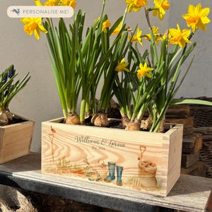 May include: A wooden planter box with a personalised inscription that reads "William & Lorna Est. 2010". The planter is filled with yellow daffodils and has a garden-themed design with blue rubber boots, gardening tools, and a sack of potatoes.