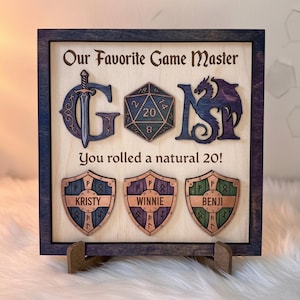 Può includere: Cartello in legno con la scritta "Our Favorite Game Master" e le lettere "GOMS" in un font fantasy. Sotto le lettere c'è la scritta "You rolled a natural 20!". Sotto ci sono tre scudi con nomi.